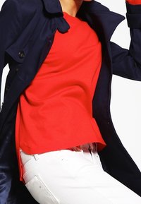 Person klädd i en ljusröd tröja, vita byxor och en mörk marinblå kappa, fångad i en dynamisk pose mot en vit bakgrund.