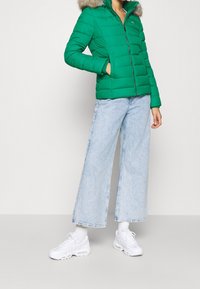 Chaqueta acolchada verde con cierre frontal, con capucha de piel sintética, combinada con jeans de pierna ancha de color azul claro y zapatillas blancas.