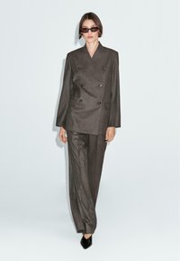 Grau taillierter, doppeltknöpfbarer Blazer mit Schulterpolstern, kombiniert mit lockeren grauen Hosen; das Modell trägt eine dunkle Sonnenbrille und schwarze spitze Schuhe.