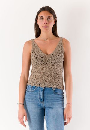 Donna con lunghi capelli scuri che indossa un top smanicato beige a maglia e jeans blu, in piedi davanti a uno sfondo bianco uniforme.