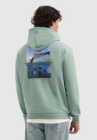 Man met krullend haar in een groene hoodie met een kleurrijke autokantoor afbeelding en de tekst "WASTELAND" op de achterkant.