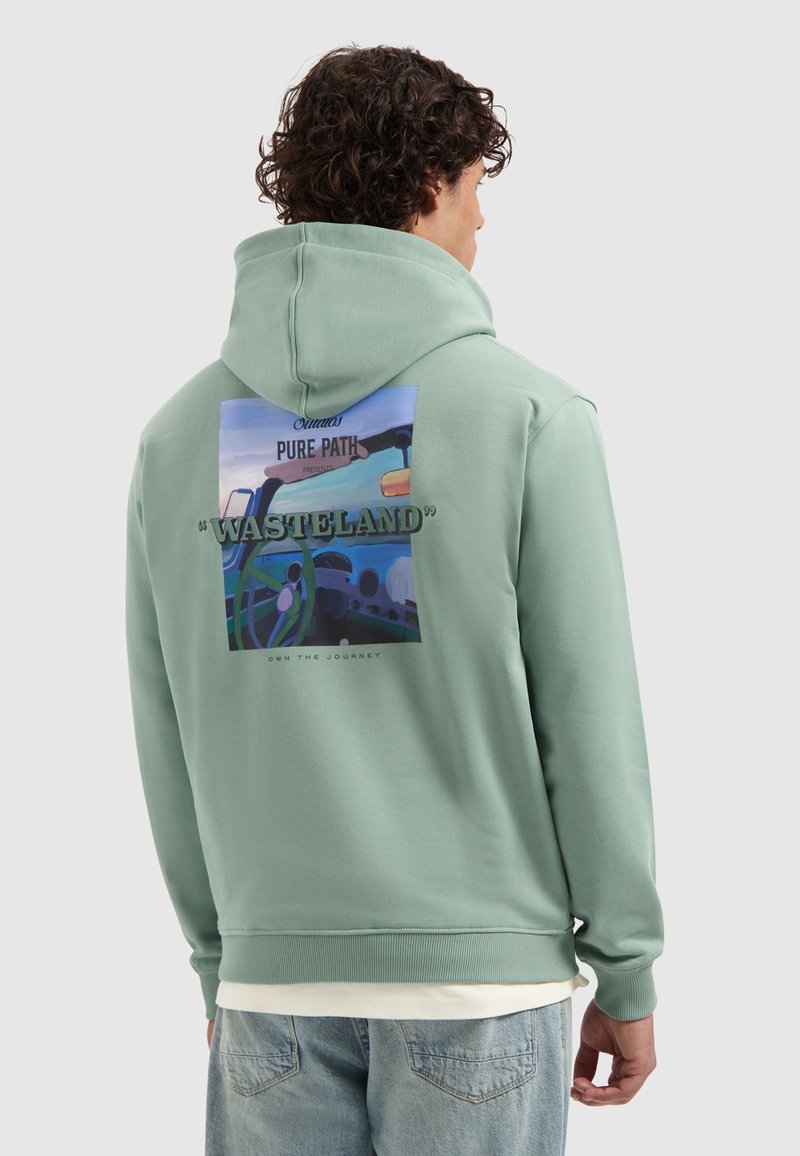 Man met krullend haar in een groene hoodie met een kleurrijke autokantoor afbeelding en de tekst "WASTELAND" op de achterkant.