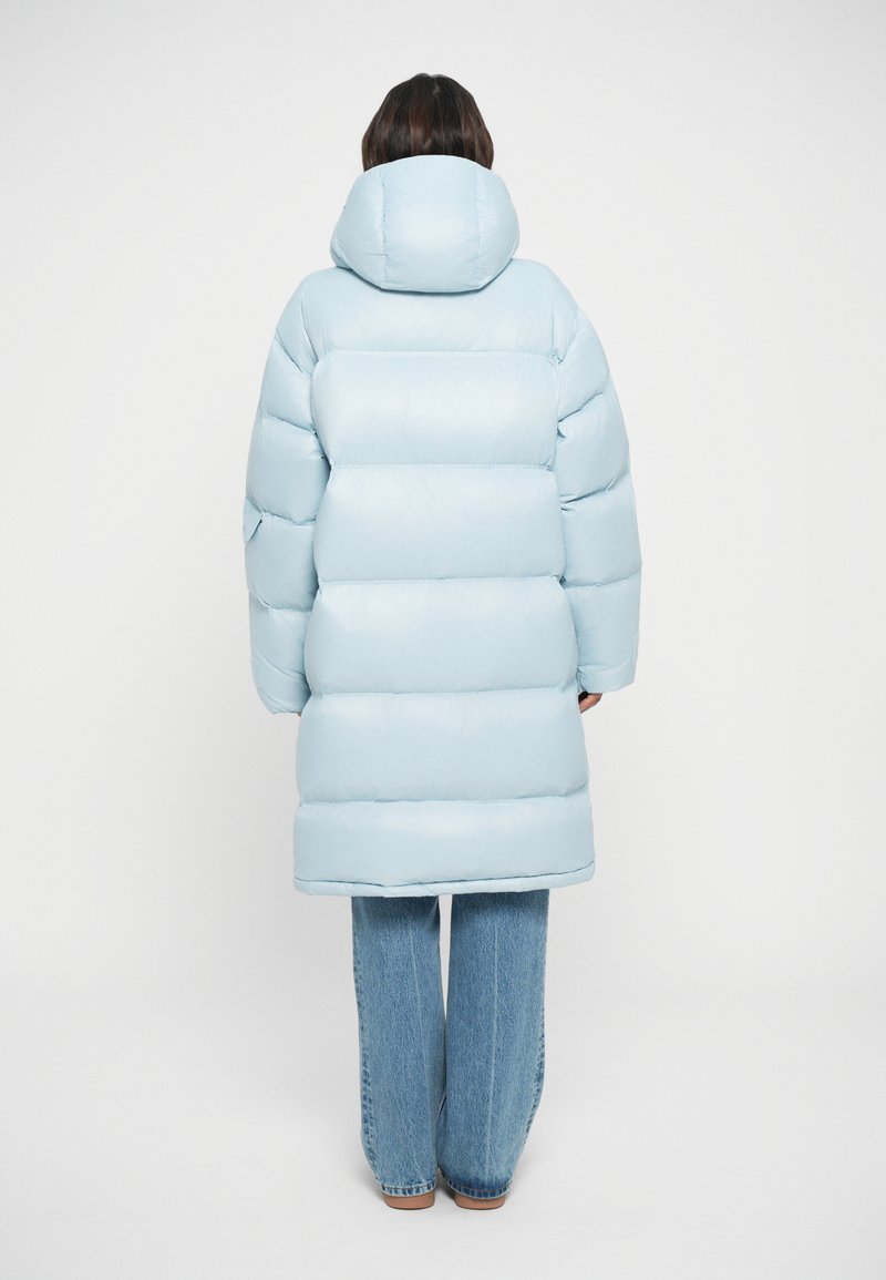 Veste en duvet bleu clair avec capuche, silhouette surdimensionnée et texture matelassée, associée à un jean en denim ample et des chaussures marron.