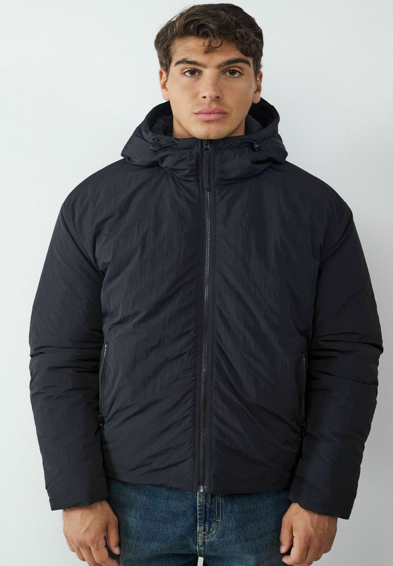 Schwarze Steppjacke mit Kapuze, vorne mit Reißverschluss, Seitentaschen und einer strukturierten Oberfläche. Getragen über blauen Jeans.