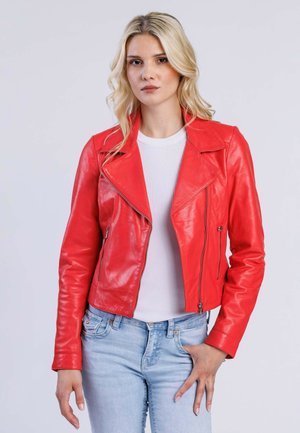 Blonde vrouw in een rode leren jas over een witte shirt en lichtblauwe spijkerbroeken, staande met één hand in haar zak tegen een effen achtergrond.