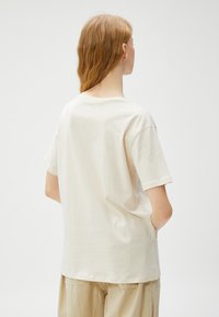 Beige bomulds t-shirt med en afslappet pasform, korte ærmer og rund halsudskæring, der har et enkelt design og blød tekstur, vist fra ryggen.