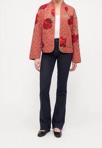 Steppjacke in Rostfarbe mit floraler Stickerei in Rot und Grün; mit langen Ärmeln, offenem Vorderteil und Seitentaschen. Kombiniert mit ausgestellten dunklen Jeans.