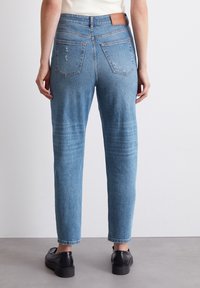 Jeans rectos de color azul claro con áreas desgastadas y bolsillos traseros. Se puede ver una etiqueta de cuero marrón claro en la cintura. Unos zapatos negros completan el atuendo.