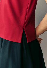 Czerwony top o gładkiej fakturze, z detalem w postaci bocznego rozcięcia. Noszony na ciemnej spódnicy, pokazujący kontrastujące kolory i warstwowy design.