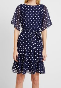 Marineblå klänning med vita prickar, genomskinliga fladdrande ärmar, midjebälte och ett flerlagers volangslut. Slät tygtextur.