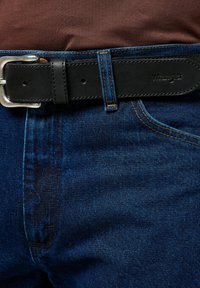 Jeans de mezclilla azul oscuro con un cinturón de cuero negro visible que presenta una hebilla de metal plateada. El logo de Wrangler está grabado en el cinturón.