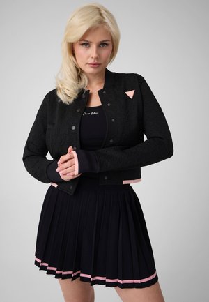 Femme blonde portant une veste noire courte boutonnée et une jupe plissée avec une bordure rose, debout les mains jointes devant un fond gris uni.