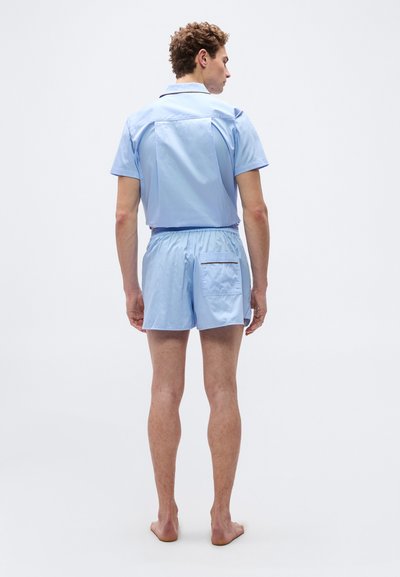 Άνδρας που στέκεται ξυπόλυτος, γυρισμένος με την πλάτη, φορώντας ανοιχτό μπλε πουκάμισο με κοντά μανίκια και ταιριαστά σορτς με πίσω τσέπη.