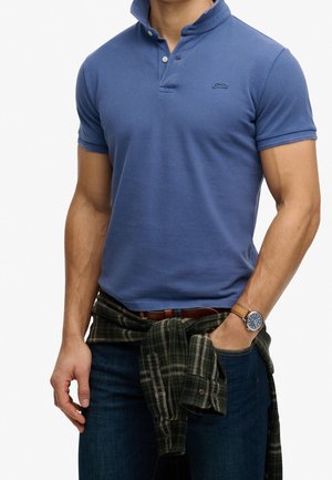 Mand iført en blå kortærmet polo, mørke jeans, en ternet skjorte bundet om livet og et armbåndsur med en brun læderrem.