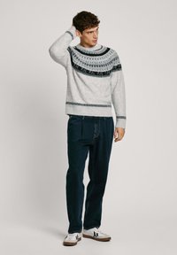Pull en tricot gris clair avec un empiècement à motifs noirs, porté avec un pantalon en denim bleu foncé et des baskets blanches avec des accents noirs.