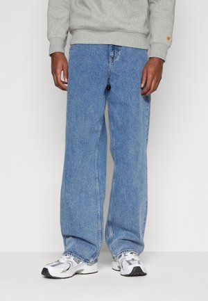 Mand iført lyseblå løstsiddende jeans, hvide og grå sneakers samt en grå sweatshirt, stående mod en ensfarvet lys baggrund.