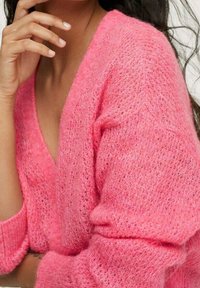 Vaaleanpunainen neulecardigan, jossa on V-pääntie, löysä tekstuuri ja joustavat hihansuut. Pehmeä, pörröinen materiaali näyttää hienot ompelutiedot.