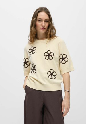 Frau mit langen, welligen Haaren, die einen cremefarbenen Kurzarm-Pullover mit braunen Blumenkonturen trägt, kombiniert mit dunkelbraunen Hosen und einem silbernen Armband.