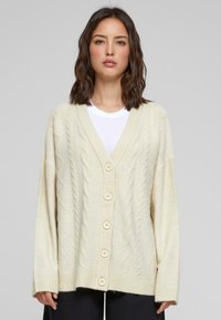 Cremfarbener Kabelstrickcardigan mit V-Ausschnitt, ausgestattet mit fünf großen beigen Knöpfen, abfallenden Schultern sowie gerippten Ärmelbündchen und Saum.
