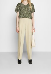 Blouse verte à motif floral avec un col en V, associée à un pantalon beige taille haute. Le mannequin tient un petit sac à main crème texturé. Chaussures noires.