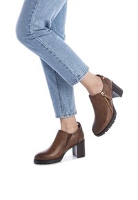 Botas de cuero marrón hasta el tobillo con un tacón grueso, detalle de cremallera lateral y suela texturizada, combinadas con jeans azules arremangados.