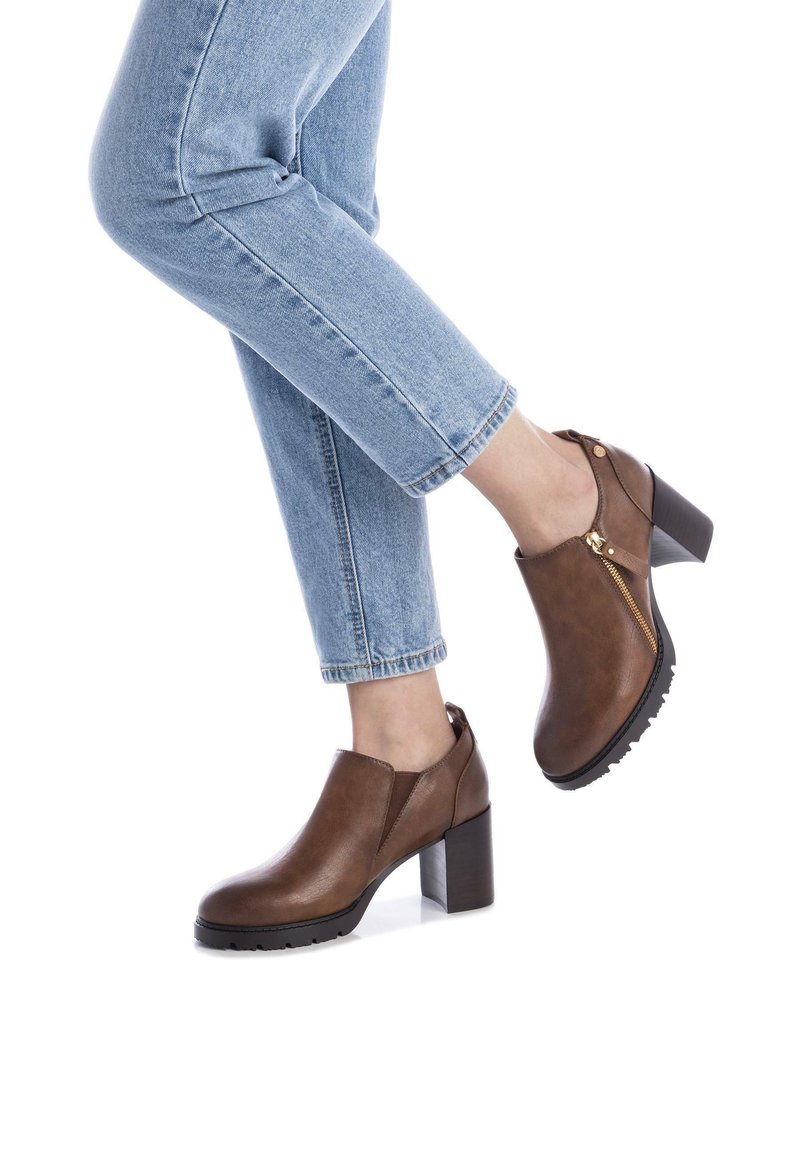 Botas de cuero marrón hasta el tobillo con un tacón grueso, detalle de cremallera lateral y suela texturizada, combinadas con jeans azules arremangados.