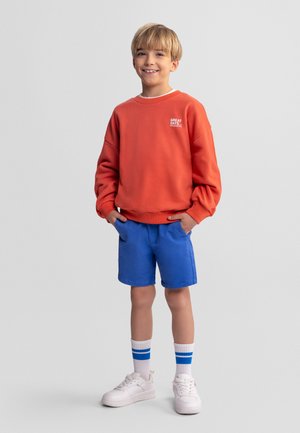 Smilende dreng med blondt hår iført orange sweatshirt, blå shorts, hvide sneakers og hvide sokker med blå striber, stående med hænderne i lommerne.