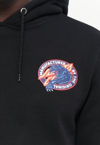 Zwarte hoodie met een ronde geborduurde patch met een blauw panterontwerp, rode en witte tekst, en een textuurkoord.
