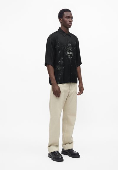 Homme portant une chemise noire à manches courtes avec un motif graphique, un pantalon beige et des chaussures noires, debout devant un fond blanc uni.
