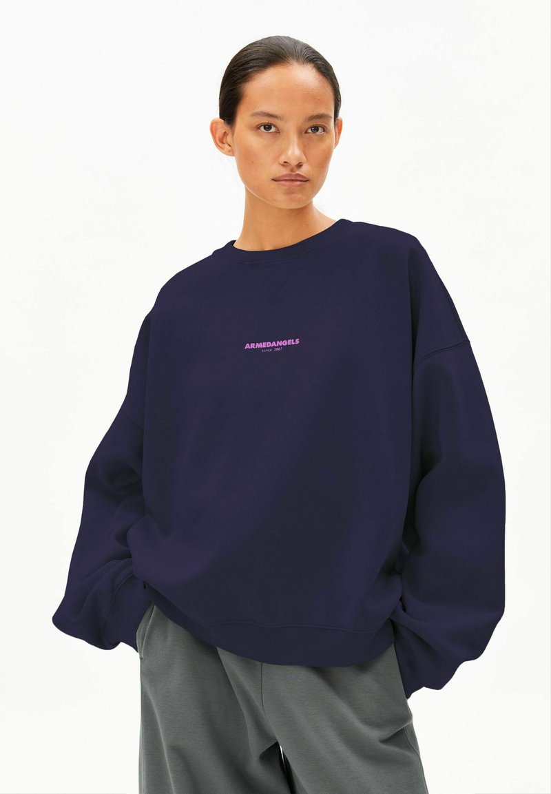 Marineblauer übergroßer Pullover mit rundem Ausschnitt und gerippten Bündchen, mit pinkem Text "ARMEDANGELS seit 2012" in der Mitte auf der Brust.