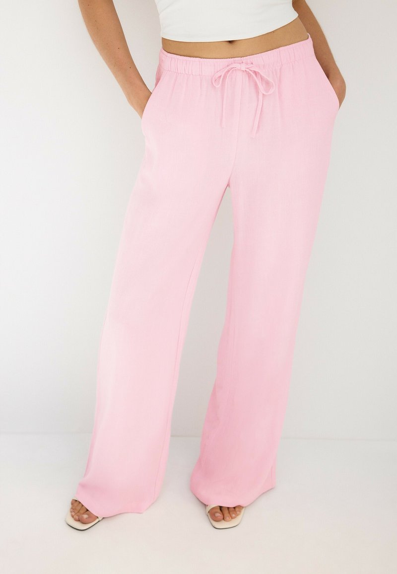 Femme portant un pantalon large rose clair avec une taille élastique et un cordon, associé à des sandales blanches et un top court blanc.