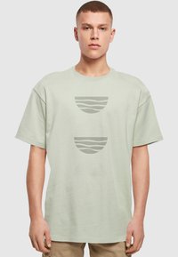 Lys grøn bomulds T-shirt med løs pasform, der har tre cirkulære grafiske designs i afdæmpet grå, herunder bølgede og stribede mønstre.