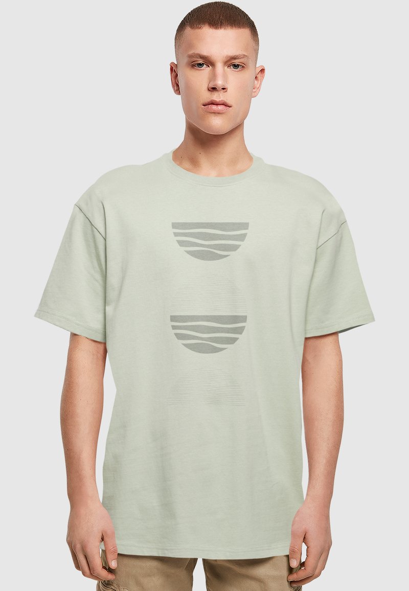 Lys grøn bomulds T-shirt med løs pasform, der har tre cirkulære grafiske designs i afdæmpet grå, herunder bølgede og stribede mønstre.