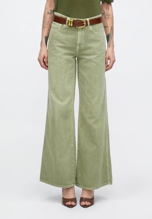 Rohelised laia säärega teksad, valmistatud denimist, millel on pruun nahkvöö kuldse pandlaga, sile tekstuur ja toored ääred.