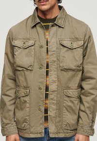 Veste utilitaire kaki avec un devant à boutons, quatre poches avant et une texture côtelée. Portée par-dessus une chemise rayée verte et orange.