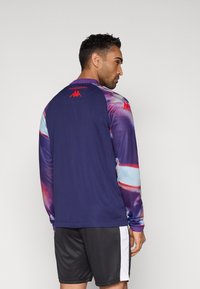 Hosszú ujjú atlétikai ing sötétkék testtel és színes, mintás ujjakal. Vörös logó található a hátoldalán, sima textúrával.