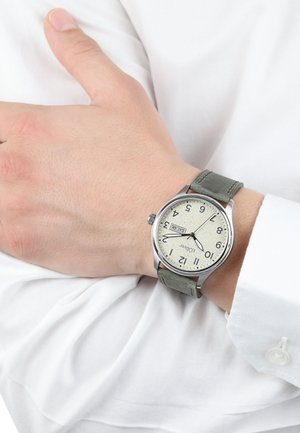 Silberne Armbanduhr mit einem hellen, strukturierten Ziffernblatt, grünem Lederarmband und arabischen Ziffern. An einer Hand getragen, die ein weißes Hemd hält.
