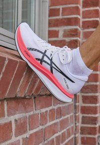 Vit löparsko med en rosa och svart sula, som har ett andningsbart mesh-material och randiga dekorativa detaljer, märkta "MAGIC SPEED."