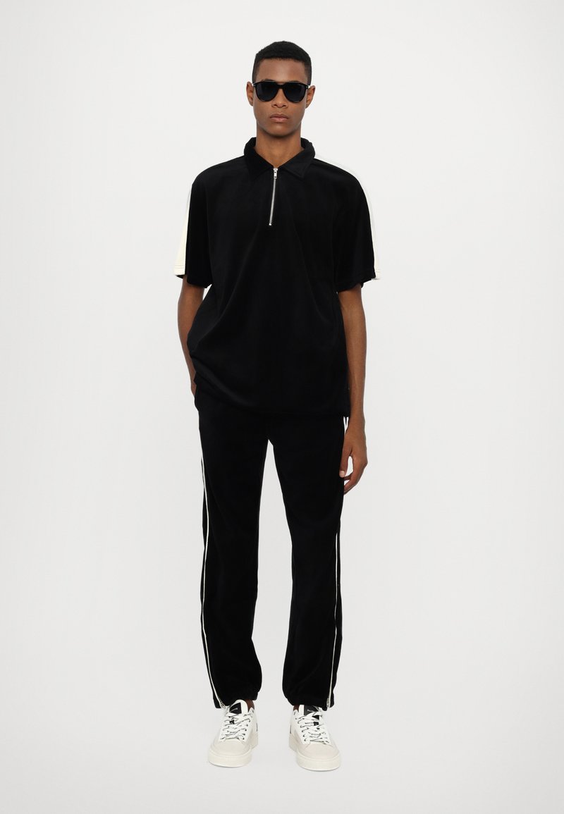 Polo en velours noir avec un col zippé, associé à un jogging noir assorti avec des rayures blanches sur les côtés, et des baskets blanches. Les accessoires comprennent des lunettes de soleil.