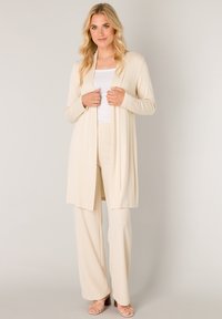 Lange crème cardigan gemaakt van zachte stof, met een open voorkant, lange mouwen en een vloeiende pasvorm. Gecombineerd met bijpassende crème broek.