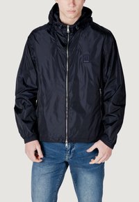 Giacca leggera navy con cappuccio, frontale con zip, polsini elastici e logo a patch sul petto. Finitura in tessuto liscia e lucida.