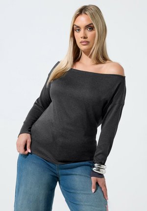 Kobieta z długimi blond włosami, ubrana w ciemny top odkrywający ramiona, niebieskie dżinsy i srebrne bransoletki, pozująca na jasnym tle.
