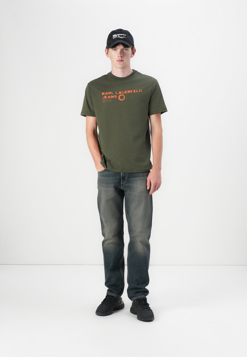 Green cotton t-shirt with orange logo, paired with faded blue jeans and black sneakers. Model wearing a black cap. Simple, casual outfit.