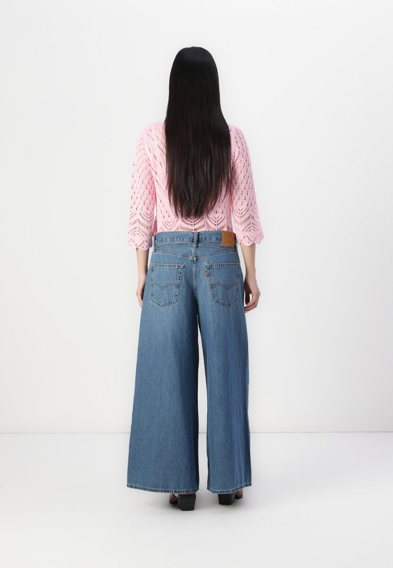 Jean en denim bleu à jambe large avec une taille haute, présentant une patch en cuir marron, deux poches arrière et une coupe décontractée.