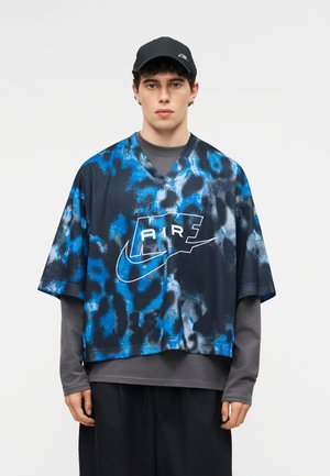 Νέος άνδρας με μαύρο καπέλο Nike, μπλε-μαύρο πουκάμισο Nike Air με σχέδιο tie-dye πάνω από γκρι μακρυμάνικη μπλούζα και άνετο μαύρο παντελόνι.