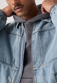 Σακάκι από ανοιχτό μπλε τζιν με φερμουάρ, φορεμένο πάνω από γκρι φούτερ με κουκούλα. Διαθέτει ορατές ραφές και ριμπ μανσέτες.