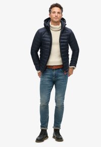 Mørkeblå dunjakke med strikket hætte, beige turtleneck, blå jeans og sorte snørebåndsstøvler. Har lynlås foran og diskret polstring.