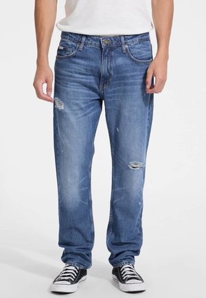 Pantalones vaqueros de mezclilla azul con un corte relajado, detalles desgastados en las rodillas, cinco bolsillos y un cierre de botón y cremallera, combinados con zapatillas negras.