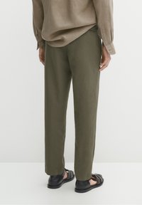 Personne portant un pantalon droit vert olive, une chemise à manches longues beige et des sandales noires à bout ouvert, debout devant un fond blanc.