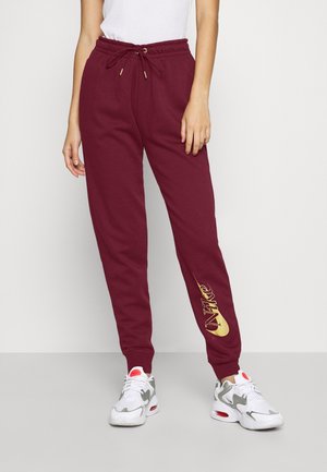 Personne portant un pantalon de jogging Nike bordeaux avec un logo doré sur la jambe gauche et des baskets blanches avec des touches rouges, debout devant un fond uni.